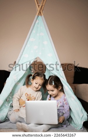 Two girls sisters watching on laptop at wigwam tent. Technology and home concept.