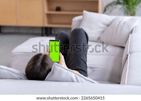 Feminine Hand Scrolling Feed on Smartphone with Green Screen Mock Up Display. Female is Relaxing on Sofa at Home, Watching Videos and Reading Social Media Posts on Mobile Device.