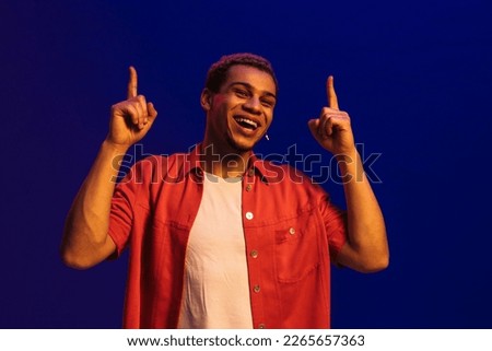 Stylish smiling young afro man pointing fingers up at copy space isolated over blue neon studio background