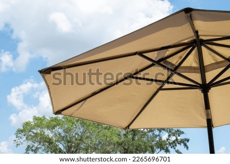 A large summer nylon patio shade umbrella, beige in color, opened with brown wooden supports. The background is a bright blue sky with white fluffy and a portion of a large tree with green leaves. 