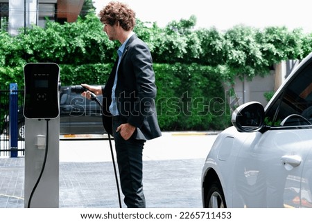 Progressive businessman with electric car recharging at public charging station at modern city residential area. Eco friendly rechargeable car powered by alternative clean energy.