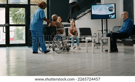 Mother and child visiting old woman in wheelchair at medical facility, talking to senior patient with disability. Little girl bringing flowers to grandmother in hospital reception. Tripod shot.