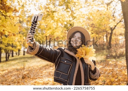 Stylish young smiling woman walking in autumn park taking selfie pictures with leaves using smartphone on tripod, wearing hat, fashion style trend. Copy space