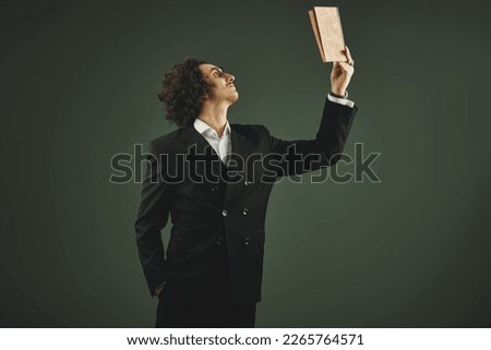A strict professor in a classic black suit and glasses stands with an open book in his hands. Studio portrait on a gray background. Scientist, teacher. Royalty-Free Stock Photo #2265764571
