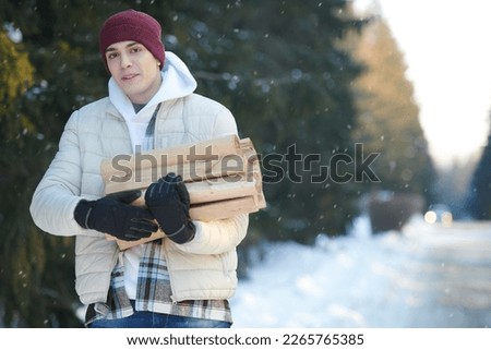 A young man in warm winter clothes holds an armful of firewood, standing in a winter snowy forest, and smiles joyfully. Active recreation in winter, travel and tourism.