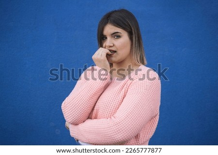 Young Caucasian plus-size woman biting her nails while looking at the camera with a demure expression. Concept of body shaming and nail-biting