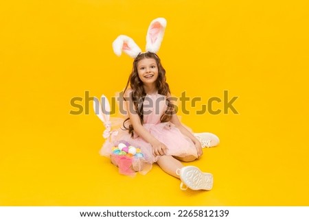 A little girl dressed as an Easter bunny with a basket of colorful eggs on a yellow isolated background. Festive spring Easter.