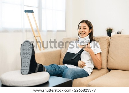 Happy woman recovery from accident fracture broken bone injury with leg splints in cast neck splints collar arm splints sling support arm in living room. Social security and health insurance concept. Royalty-Free Stock Photo #2265823379