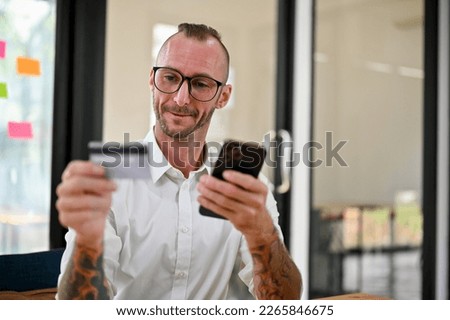 Happy Caucasian man with glasses and tattoo on his arms sits at his desk holding a credit card or debit card and smartphone, using mobile banking to pay bills or transfer money.