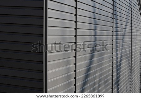 gray building is lined with square panels. metal sheet wall cladding with scalloped design. corrugated sheet, dark gray, riveted. horizontal stripes, shadows, corner, fence, aluminium, sheet