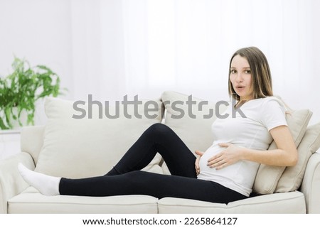 Facial expression of pregnant woman on white sofa.
