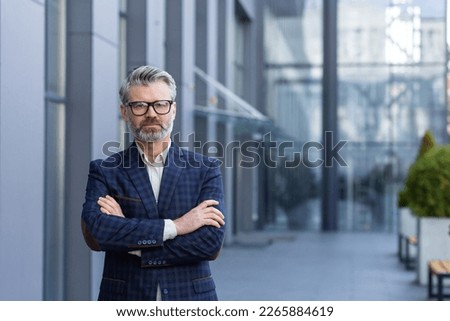 Portrait of mature successful boss, gray haired businessman looking at camera with crossed arms, man outside office building in business suit and glasses, satisfied and focused banker investor. Royalty-Free Stock Photo #2265884619