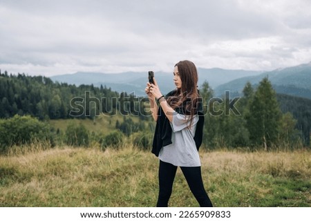 Attractive female tourist in the mountains takes a photo on a smartphone camera and looks away against the background of a beautiful landscape. Tourism concept.