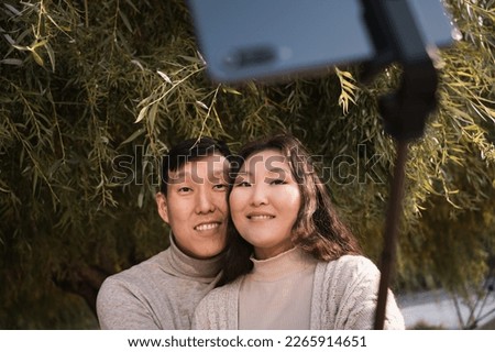 Happy young adult asian couple in love taking selfie photo by phone under big green tree of weeping willow in summer
