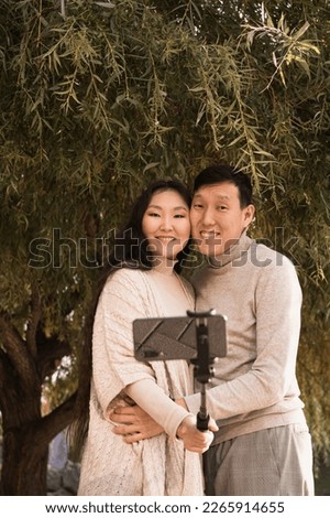 Happy hugging young asian couple in love taking selfie photo by phone under big green tree at summer day. Vertical portrait