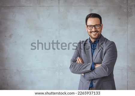 Portrait of happy businessman with arms crossed standing in office. Portrait of young happy businessman wearing grey suit and blue shirt standing in his office and smiling with arms crossed