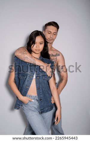 shirtless tattooed man embracing asian woman posing with hand in pocket of jeans on grey background