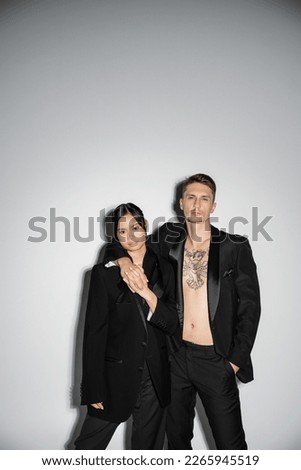 tattooed man in black suit embracing elegant asian woman while posing on grey background