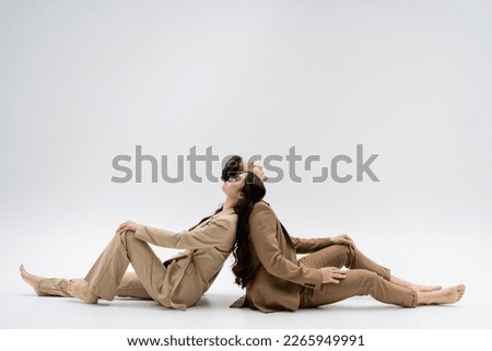 side view of multiethnic couple in beige suits sitting back to back on grey background Royalty-Free Stock Photo #2265949991