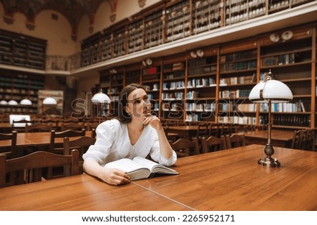 Dreamy beautiful woman sitting in a public library with a book and looks away. Studying in the library