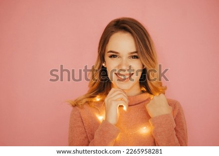 Portrait of happy lady in pink clothes and christmas lights isolvoan on pink background, looking at camera with smile on face. Christmas and New Year.