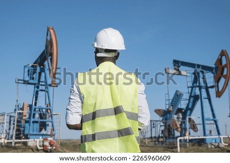 African American man controls beam-balanced pumping unit operation on sunny meadow. Black developer wearing working clothes explores production of oil