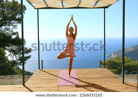 Sporty young woman practicing yoga on the wooden terrace with beautiful ocean view. Fit yogini doing the tree pose. Mountain and sea background, copy space.
