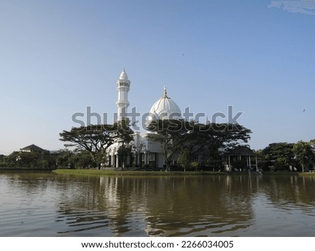 Wide view of a beautiful white mosque surrounded by big trees an a big lake.