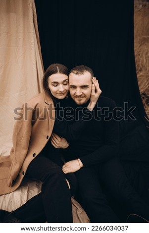 a man and a girl in black clothes and a beige coat on a black and beige fabric background. man and woman hugging, cold cloudy weather and dry yellow tall grass . stylish fashion portrait.
