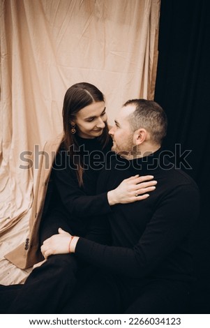 a man and a girl in black clothes and a beige coat on a black and beige fabric background. man and woman hugging, cold cloudy weather and dry yellow tall grass . stylish fashion portrait.