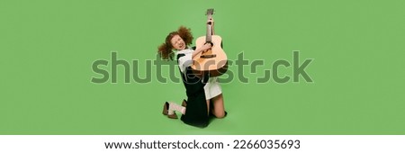 Beautiful little girl, funny musician wearing huge oversize clothes playing guitar, having fun isolated over green background. Pop art, music, new vision, fun concept. Banner, flyer for ad