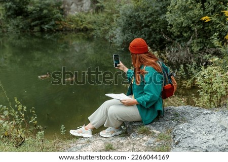 Beautiful female in red hat hiking during free time at national park. View from back. Happy woman enjoying her holidays on wild nature. Hiker taking photo with smart phone