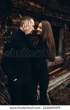 a man and a woman in black clothes near an old wooden house. photo in dark brown tones. old abandoned house and cloudy cold weather. beautiful stylish couple on the background of a brown wooden wall.