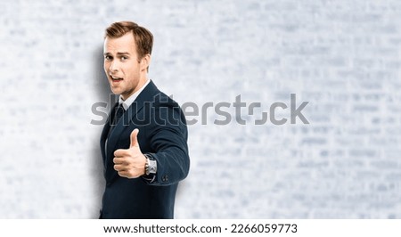 Excited businessman in black suit showing thumbs up like hand sign gesture, in blue confident suit, on white bricks wall background. Handsome happy man. Copy space for ad, slogan.