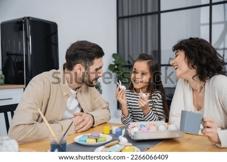 Smiling parents looking at happy daughter with Easter rabbits near eggs and macaroons at home