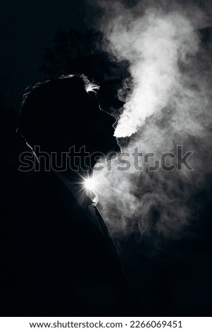 Dark photo of a silhouette of a man blowing smoke against the light. Black background and white smoke. Night portrait of a smoking man