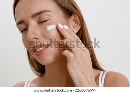 Close up face portrait of young woman applying cream to her face on white studio background