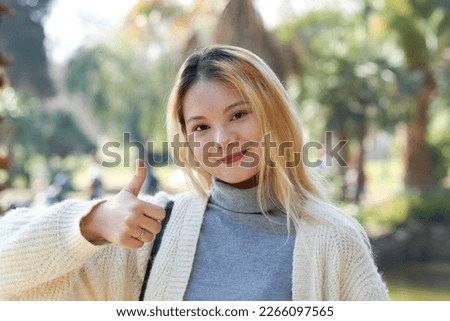 Asian young girl shows thumb up looking at the camera in a park. Positive girl makes a sign of approval or ok.