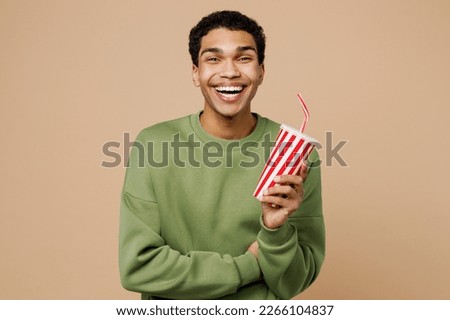 Young fun man of African American ethnicity wear green sweatshirt hold in hand cup of soda cola fizzy water look camera isolated on plain pastel light beige background studio. People lifestyle concept Royalty-Free Stock Photo #2266104837
