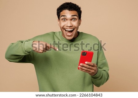 Young fun surprised man of African American ethnicity wearing green sweatshirt hold in hand use point index finger on mobile cell phone isolated on plain pastel light beige background studio portrait Royalty-Free Stock Photo #2266104843