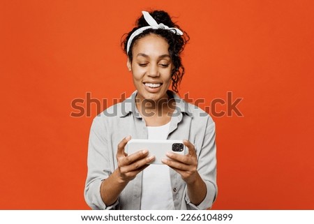 Gambling young woman of African American ethnicity she wears grey shirt headband use play racing app on mobile cell phone hold gadget smartphone for pc video games isolated on plain orange background