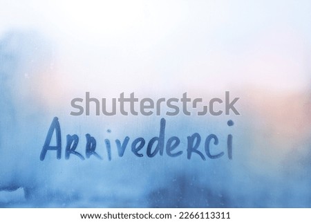 Handwritten lettering Arrivederci in Italian language is goodbye in english on misted glass on sunset blue orange window flooded with raindrops