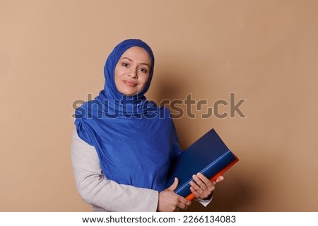 Beautiful Middle-Eastern Muslim woman with head covered in a blue hijab, confident female educator teacher professor, holding books and looking at camera, on isolated beige background. Erudite people
