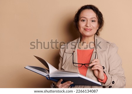 Beautiful happy middle aged brunette woman in casual wear, holding eyeglasses and smiling, looking at camera, isolated on beige background, reading a book. The concept of erudite educated people