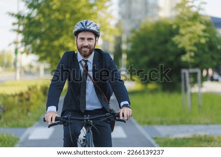Smiling male employee in black suit commuting to work on bike lane in sunny morning. Front view of happy bearded worker looking at camera, while driving bicycle outdoors. Concept of eco lifestyle.