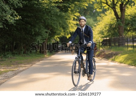 Smiling bearded executive in business apparel returning from job on bike through green area. Front view of joyful office clerk feeling great, while cycling home in sunny day. Concept of eco lifestyle. Royalty-Free Stock Photo #2266139277
