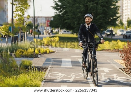 Happy office clerk in smart casual smiling, while riding after work in downtown. Front view of enjoyed entrepreneur cycling on bike lane, while returning home at sunny day. Concept of eco lifestyle. Royalty-Free Stock Photo #2266139343