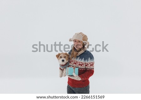 girl in a brown coat gives a Jack Russell Terrier a gift on a white background during a blizzard. The dog sits on the hands of a man. Christmas concept.