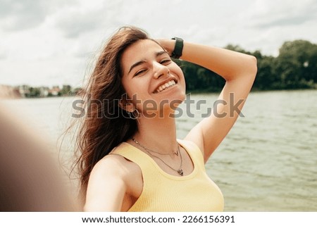 Woman standing smiling near river sea lake, take picture, make selfie hold camera. Female on beach island. Girl tourist on vacation, weekend relaxing resting resort. Lady go on shore, sunbathing

