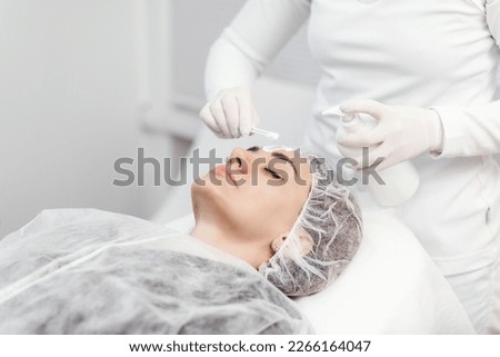 Cosmetologist is applying cream with anesthesia on patient's face skin before biorevitalization procedure. Woman in beauty clinic with doctor beautician preparing to treatment using numbing cream.